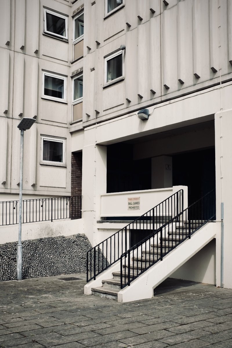 A small white sign reading 'Private Property — Ball Games Prohibited' mounted on the side of a stairwell at Blackdown Flats, a 1960s block of flats in Southend-on-Sea. Black metal railings, pebble-dash wall, paved courtyard.